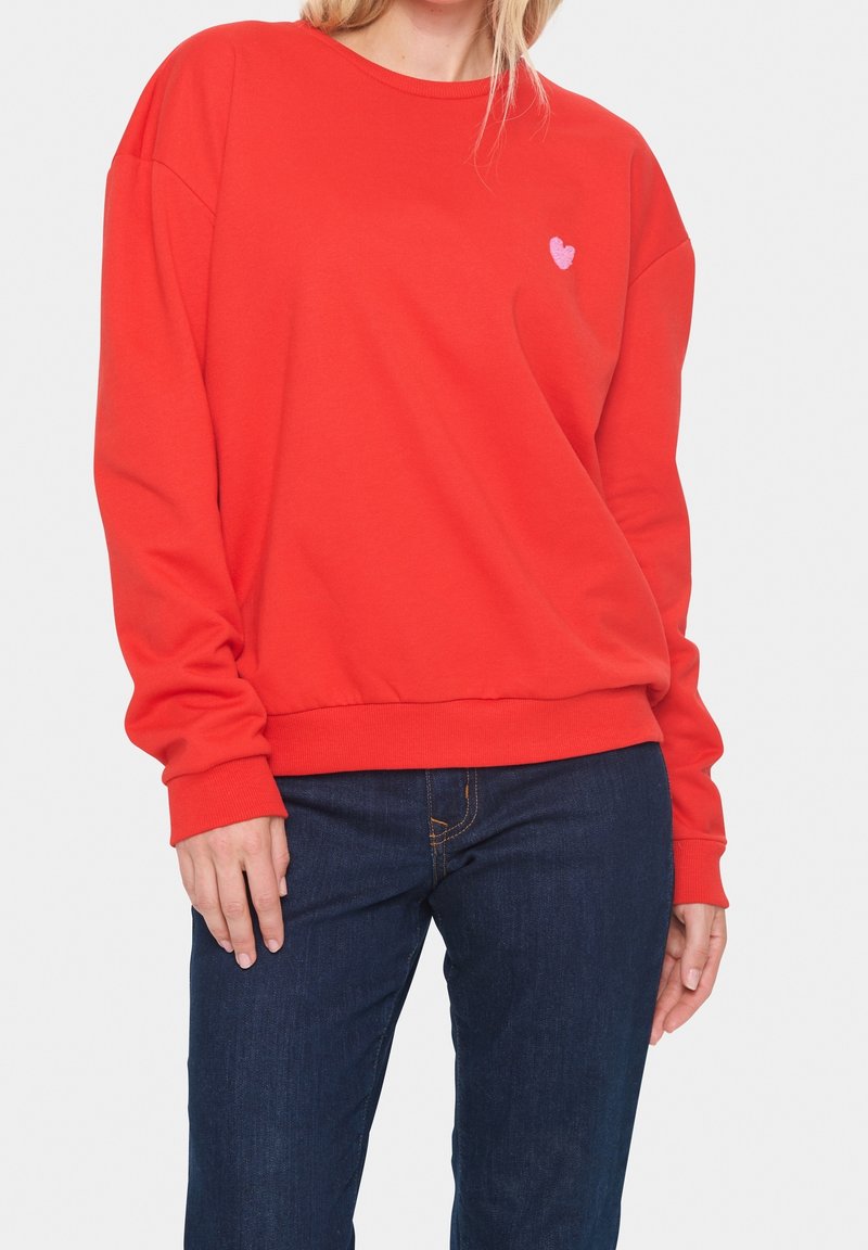 Femme portant un sweat-shirt rouge vif à manches longues avec un petit détail cœur rose sur la poitrine et un jean bleu foncé.