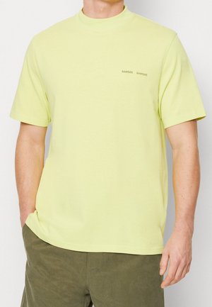 Homme portant un T-shirt à manches courtes jaune pâle avec un petit logo noir "SAMSØE SAMSØE" sur la poitrine et un pantalon vert olive, debout avec une main dans la poche.