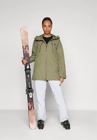 Oliwkowo-zielona kurtka narciarska na zamek z kapturem, w połączeniu z białymi spodniami. Osoba trzyma różowo-czarne narty z wiązaniami.