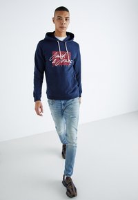 Felpa con cappuccio navy con lacci bianchi e stampa grafica, abbinata a jeans chiari strappati e sneakers scure, in piedi su uno sfondo semplice.