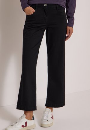 Personne portant un pantalon noir large et court, un haut violet à manches longues et des baskets blanches avec des accents bordeaux et bleu marine.