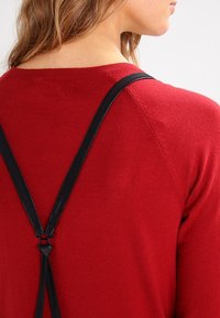 Femme portant un pull rouge avec des bretelles noires croisées dans le haut du dos, les cheveux détachés, de dos face à l'appareil photo.