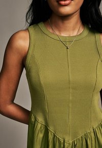 Abito verde a coste con corpetto aderente e gonna a campana, dotato di scollatura rotonda e dettagli di cucitura discreti. Collana d'argento con pendente.
