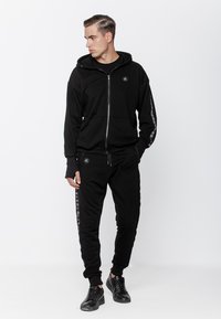 Czarny hoodie na zamek błyskawiczny z przednią kieszenią i subtelnym logo. Połączony z czarnymi dresami z bocznymi akcentami. Gładka faktura, swobodny krój.