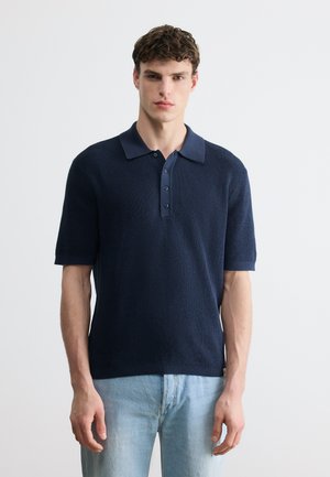 Ung mann står, iført en marineblå kortermet strikket polo og lyseblå jeans, mot en enkel hvit bakgrunn.