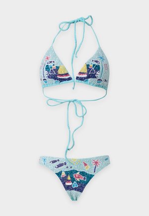 Moteris su ilgais tamsiais plaukais, vilkinti šviesiai mėlynus raštuotus bikini, stovinti basomis ant paprasto baltos spalvos fono.