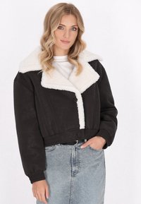 Schwarze cropped Jacke mit weißem Kunstfellkragen, Knopfverschluss und elastischen Manschetten, kombiniert mit einem hellblauen Denim-Rock.