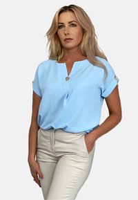 Jasnoniebieska bluzka z krótkim rękawem z dekoltem w kształcie litery V i dekoracyjnymi guzikami. Materiał ma subtelnie wyczuwalną teksturę. Zestawiona z jasno beżowymi dopasowanymi spodniami.