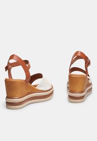Sandali con zeppa, caratterizzati da una parte superiore bianca, cinghie in cuoio marrone e un tacco dalla texture in legno. Soletta morbida con forma arrotondata e dettagli nei toni neutri.