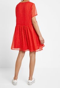Robe rouge à manches courtes en tissu léger et transparent, avec une taille froncée et de petits motifs floraux blancs. Portée avec des baskets blanches.