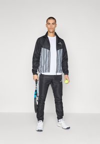 Czarna kurtka sportowa z szarymi wzorami, w połączeniu z czarnymi spodniami. Model trzyma rakietę tenisową oraz piłkę, ubrany w białe sneakersy.