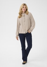 Blusa beige a maniche lunghe con colletto arricciato e chiusura a bottoni, abbinata a jeans blu scuro. Le ballerine nere completano l'outfit.