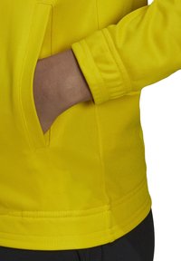 Giacca gialla con tessuto testurizzato, dotata di una tasca laterale e polsini elasticizzati, messa in risalto su uno sfondo contrastante.