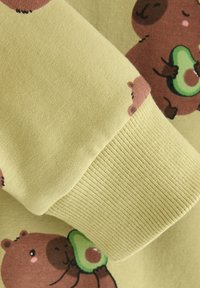 Langarmstof met een zachte gele achtergrond en cartoonbeertjes die avocado's vasthouden. De manchetten hebben een geribbelde textuur in bijpassend geel.