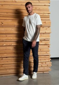 Bílý tričko s kulatým výstřihem a vyšívaným textem, navy modré slim-fit kalhoty a bílé tenisky, na pozadí dřevěných prken.