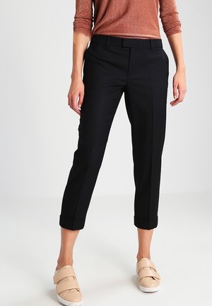 Frau trägt schwarze, schmal geschnittene, verkürzte Hosen, metallisch-braunes Langarmoberteil und beige Sneakers mit Klettverschluss, steht auf weißem Boden.