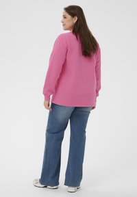 Femme aux longs cheveux bruns, portant un sweat-shirt rose, un jean bleu et des baskets blanches, se tenant debout et regardant sur le côté contre un fond uni.