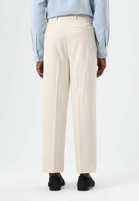 Beige trousers with a straight fit, made from smooth fabric, featuring a back pocket, and paired with black suede shoes.