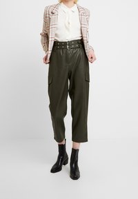 Zelené kalhoty z umělé kůže s kapsami na stranách, vysokým pasem a designem s páskem, spárované s kostkovaným blejzrem a černými kotníkovými botami.