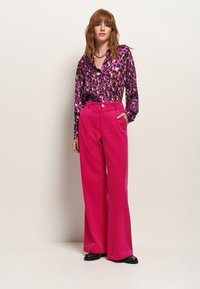 Blusa a stampa leopardata viola con maniche lunghe, abbinata a pantaloni in corduroy rosa a vita alta e ballerine nere. Dettaglio con bottone sui pantaloni.