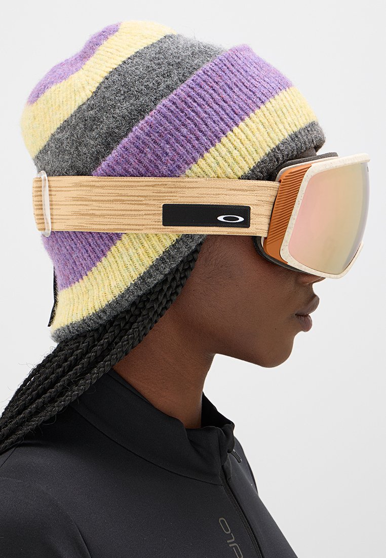 Person mit einer lila, gelben und grauen gestreiften Strickmütze, geflochtenen Haaren und reflektierenden Skibrillen mit beigem Band.