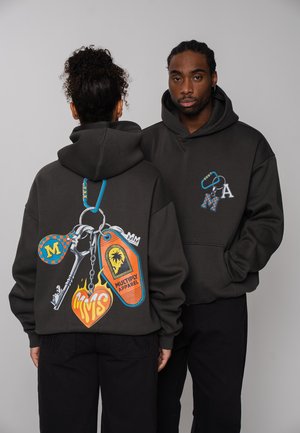 Two people wear black hoodies featuring colorful keychain designs, including keys, tags, and heart shapes with brand logos and text.