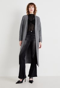 Lang grå cardigan laget av strikkemateriale, kombinert med en svart stripete bodysuit, høytidte svarte jeans og spiss svarte heler.