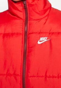 Röd isolerad pufferjacka med hög krage, framzip och vit broderad Nike-logotyp på bröstet. Slät, glänsande yta.