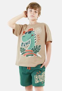 Brun t-shirt med dinosaur-grafik, iført solbriller og en kasket; parret med grønne shorts med "RAWR" tryk og orange snor.