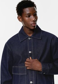 Giacca di jeans scuro con colletto alto e due tasche frontali, caratterizzata da cuciture a contrasto e chiusure a bottone. Tessuto morbido.