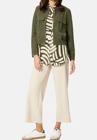 Olivgrüne kurze Jacke mit zwei Brusttaschen, darunter ein schwarz-weiß gestreiftes Hemd, cremefarbene weite Hose und weiße Turnschuhe.