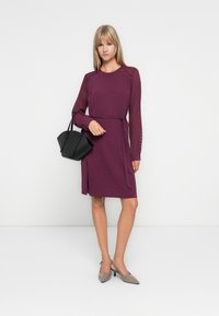 Rochie purpurie cu mâneci lungi din plasă, talie legată și croială dreptă. Asortată cu o geantă de mână neagră și tocuri gri ascuțite.