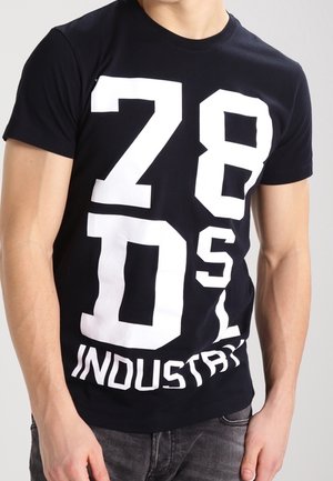 Homme portant un T-shirt noir avec de grandes lettres et chiffres blancs affichant "28 DSL INDUSTRY" sur le devant.