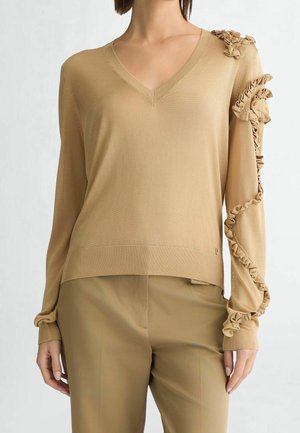 Kobieta ubrana w beżowy sweter z dekoltem w serek i teksturowaną falbanką na prawym ramieniu i rękawie, zestawiony z beżowymi spodniami.