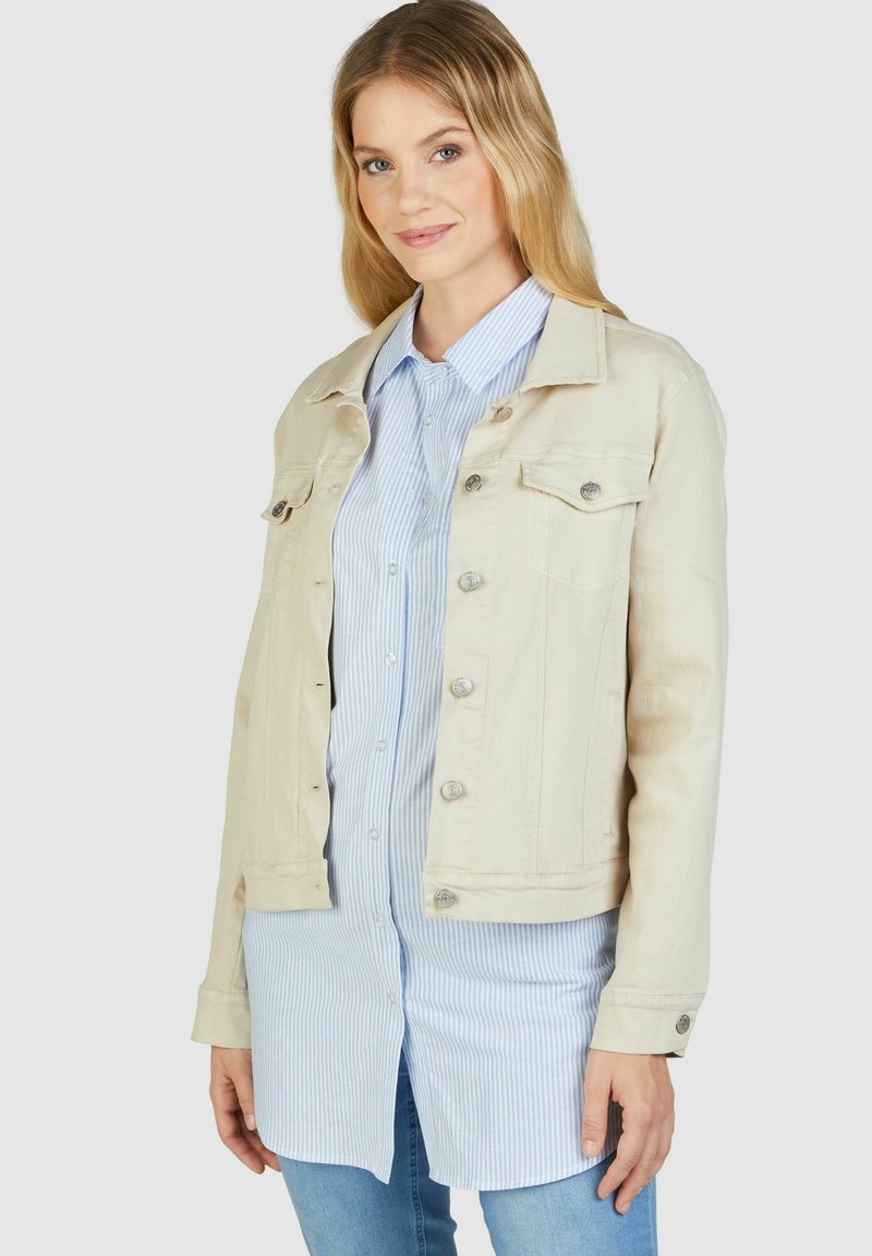 Beige Denimjacke mit zwei Brusttaschen, silbernen Druckknöpfen und taillierter Passform, getragen über einem hellblauen gestreiften Hemd.