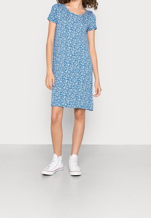 Femme portant une robe fleurie bleue à manches courtes et des baskets blanches montantes, debout sur un sol gris clair contre un mur blanc.