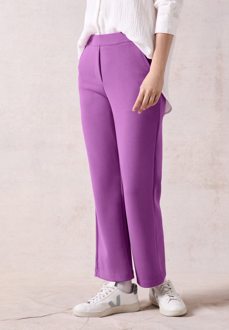 Pantalons larges violets avec une texture lisse, dotés d'une taille haute et de détails plissés. Associés à des baskets blanches avec des accents gris.