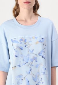 Light blue oversized t-shirt featuring a large floral print in pastel blue and beige, with a smooth, soft texture and crew neckline.