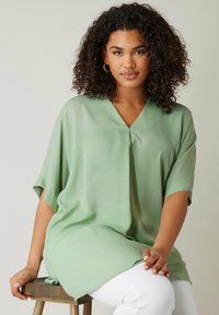 Ljusgrön, löst sittande blus med korta ärmar och V-ringning. Tillverkad av ett mjukt tyg, har en lätt drapering och sidoflikar.