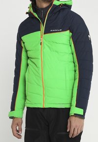 Homme portant une veste matelassée verte et bleu marine avec une fermeture éclair orange et des détails orange, assortie à un pantalon noir.