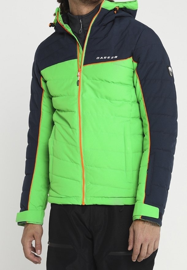 Homme portant une veste matelassée verte et bleu marine avec une fermeture éclair orange et des détails orange, assortie à un pantalon noir.