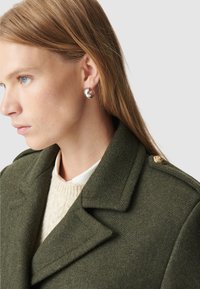 Cappotto in lana verde oliva con colletto strutturato, dotato di un bottone dorato. Orecchini a cerchio argentati adornano l'orecchio della modella. Maglione bianco sotto.