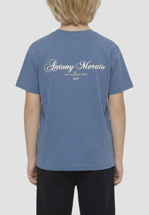 Bambino con capelli biondi che indossa una maglietta blu Antony Morato con logo bianco e pantaloni neri, mostrato di spalle su sfondo semplice.
