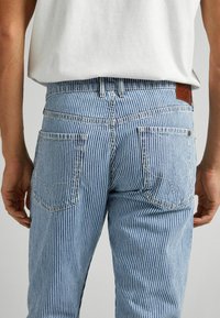 Randiga denimjeans i ljusblått med vita vertikala ränder, utrustade med bakfickor och en brun läderlapp på midjebandet.