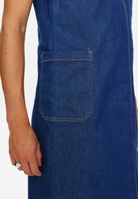 Denim obleka v temno modri barvi, z bočno žepom in kontrastnim šivanjem ter teksturirano tkanino, ki poudarja sproščen oblikovanje oblačila.