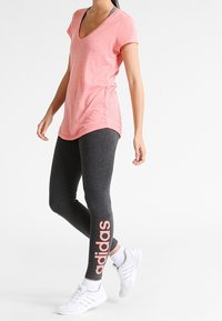 Różowa koszulka typu v-neck oraz grafitowe legginsy z różowym logo "adidas". Białe sneakersy dopełniają sportowy strój. Gładka faktura materiału.