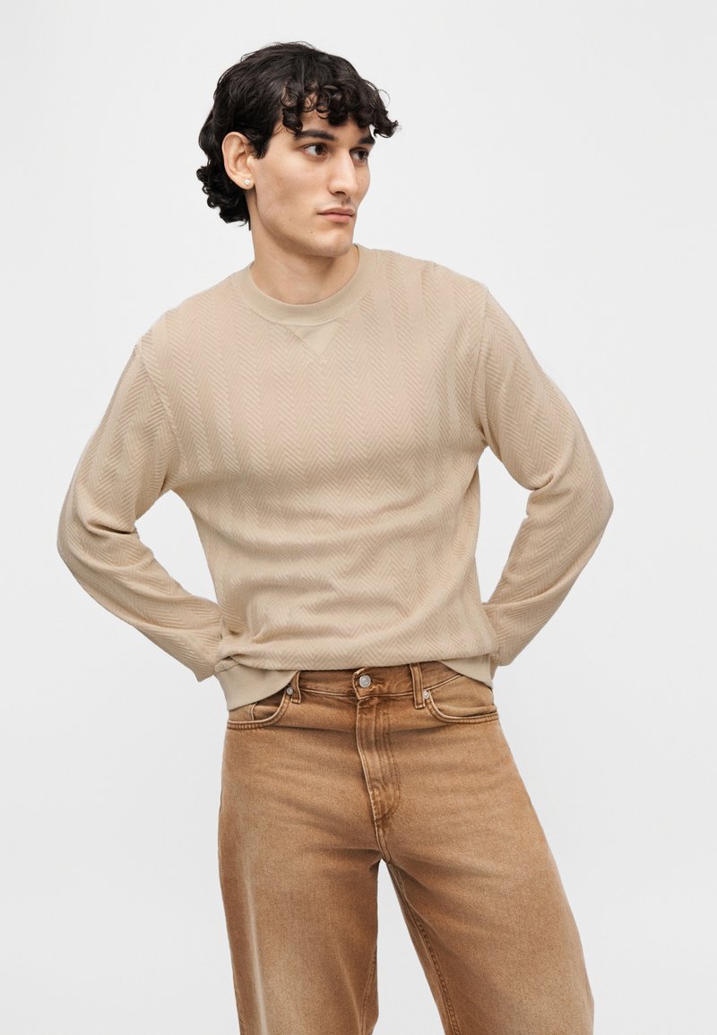 Pull en maille beige texturée avec un motif chevron, col rond et poignets côtelés, associé à un jean marron clair.
