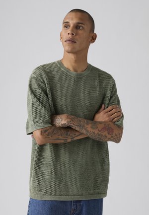 Hombre con los brazos cruzados mostrando tatuajes, vistiendo un jersey verde de manga corta con textura y vaqueros azules sobre un fondo liso.