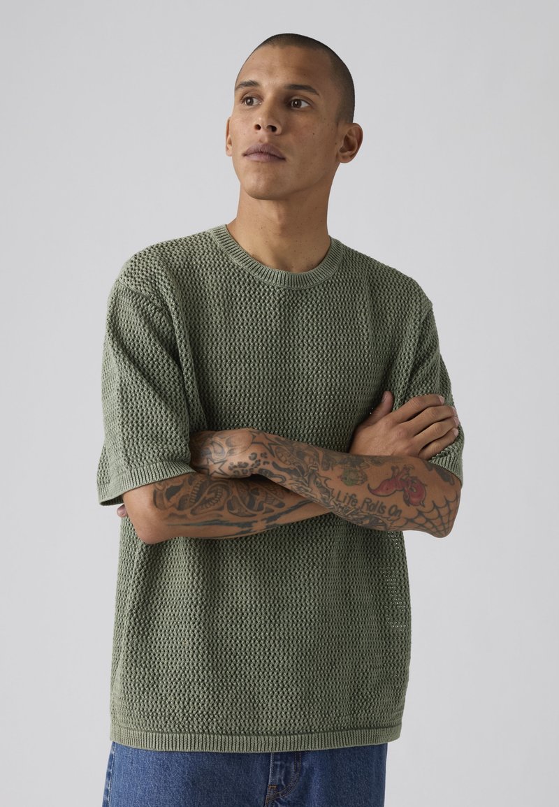 Hombre con los brazos cruzados mostrando tatuajes, vistiendo un jersey verde de manga corta con textura y vaqueros azules sobre un fondo liso.