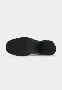 Semelle d'une chaussure habillée en cuir noir avec bout carré et détail de boucle, vue par le dessous sur fond clair uni.